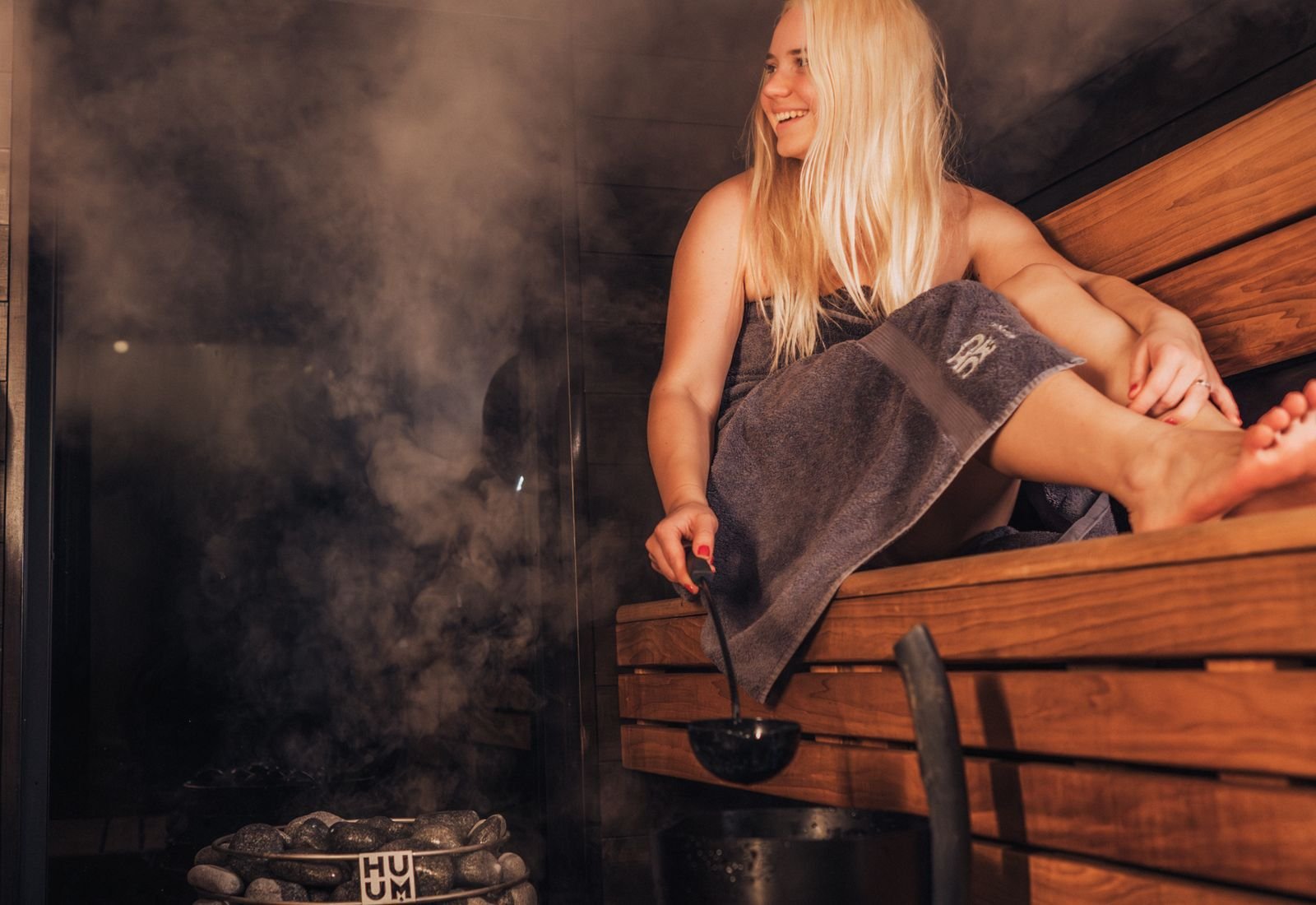 Woman wrapped in a towel on a wooden sauna bench with steam rising from the stove